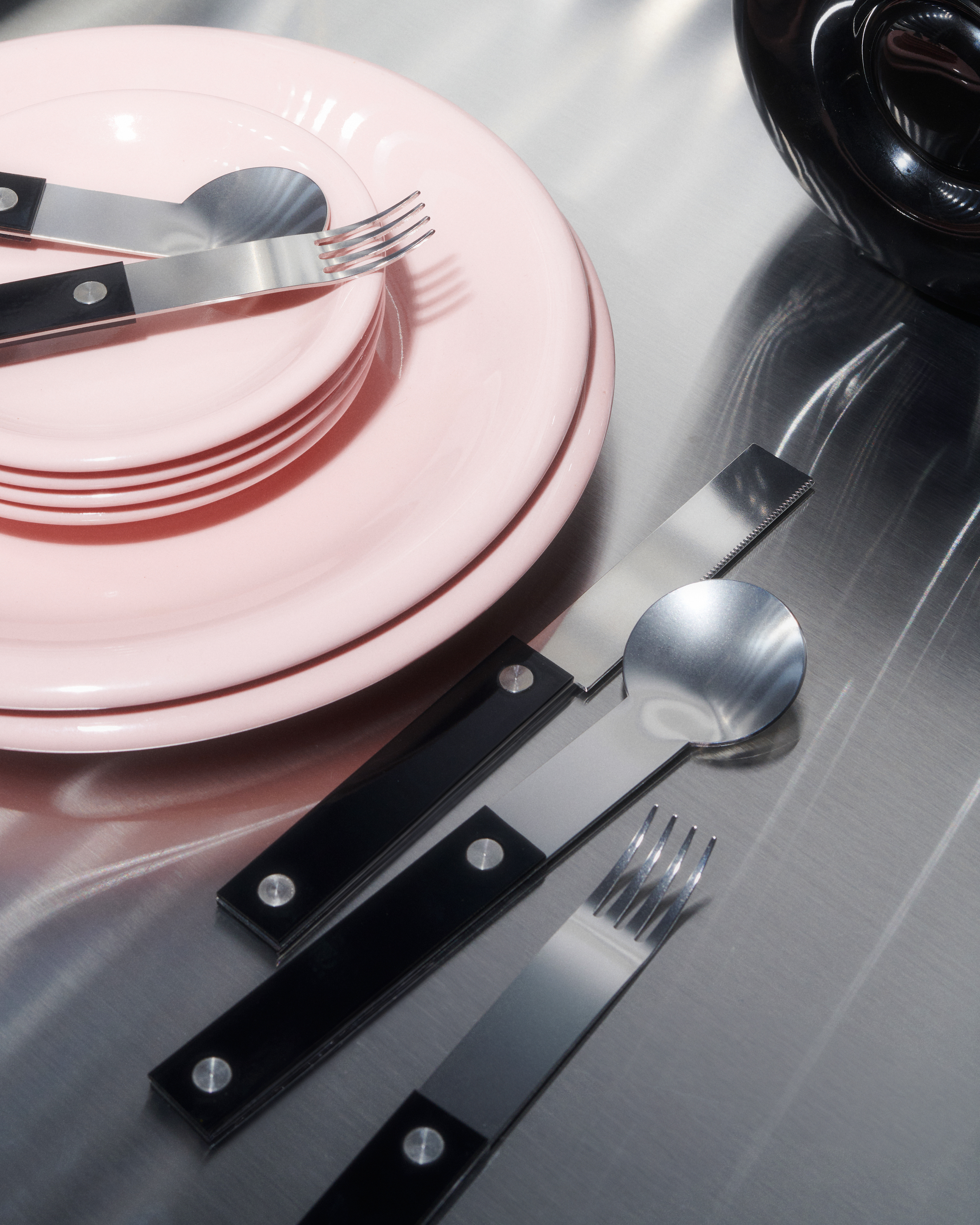 forks-plus-flatware-black-handles-pink-plates-steel-surface Stainless steel flatware with black handles beside pink plates on a stainless steel surface