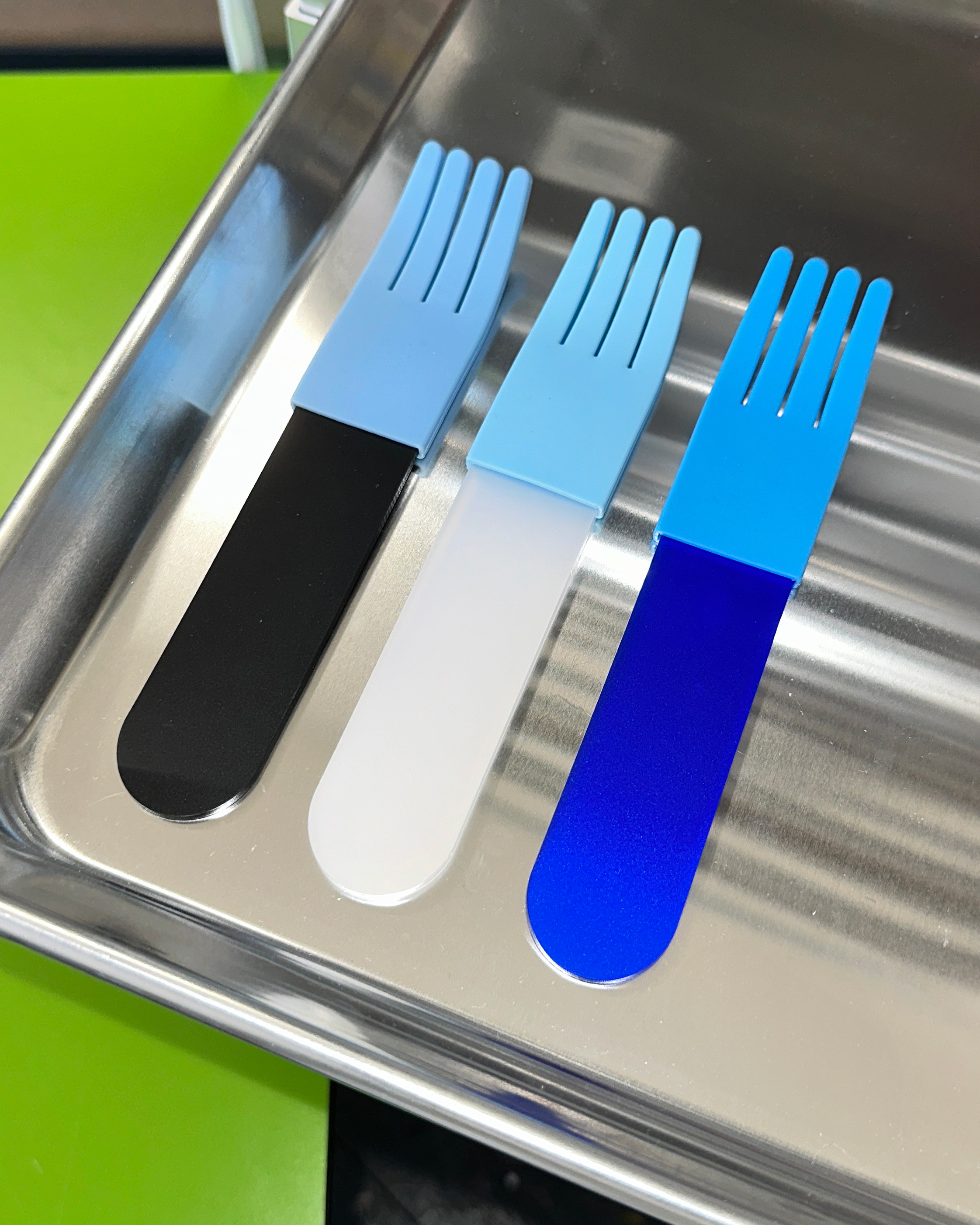 forks-plus-blue-acrylic-fork-trio-stainless-steel-tray Trio of blue acrylic forks sitting on stainless steel tray with green background surface